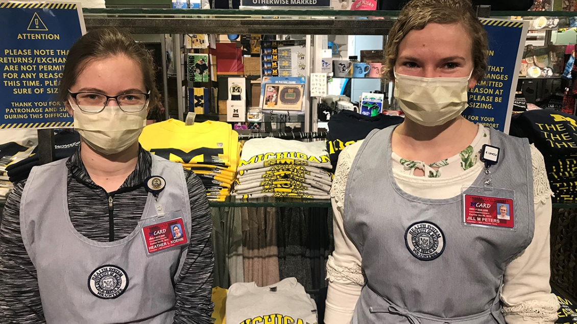 Two female Gift Shop volunteers wearing masks and gray volunteer uniforms standing in front of stacks of t-shirts